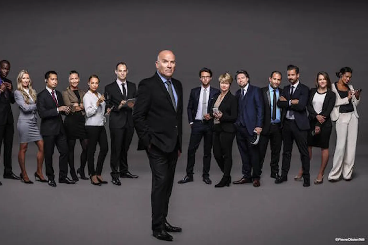 Des candidats participent à une épreuve collective dans un bureau, sous le regard d’un dirigeant et de caméras de télévision.